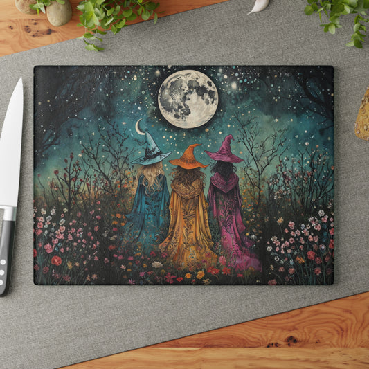 Decorative cutting board with three witches in colorful hats against a night sky with a moon, on a gray surface.